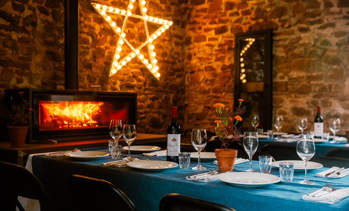 Table laid for dining in stone barn with fire blazing in wood burner